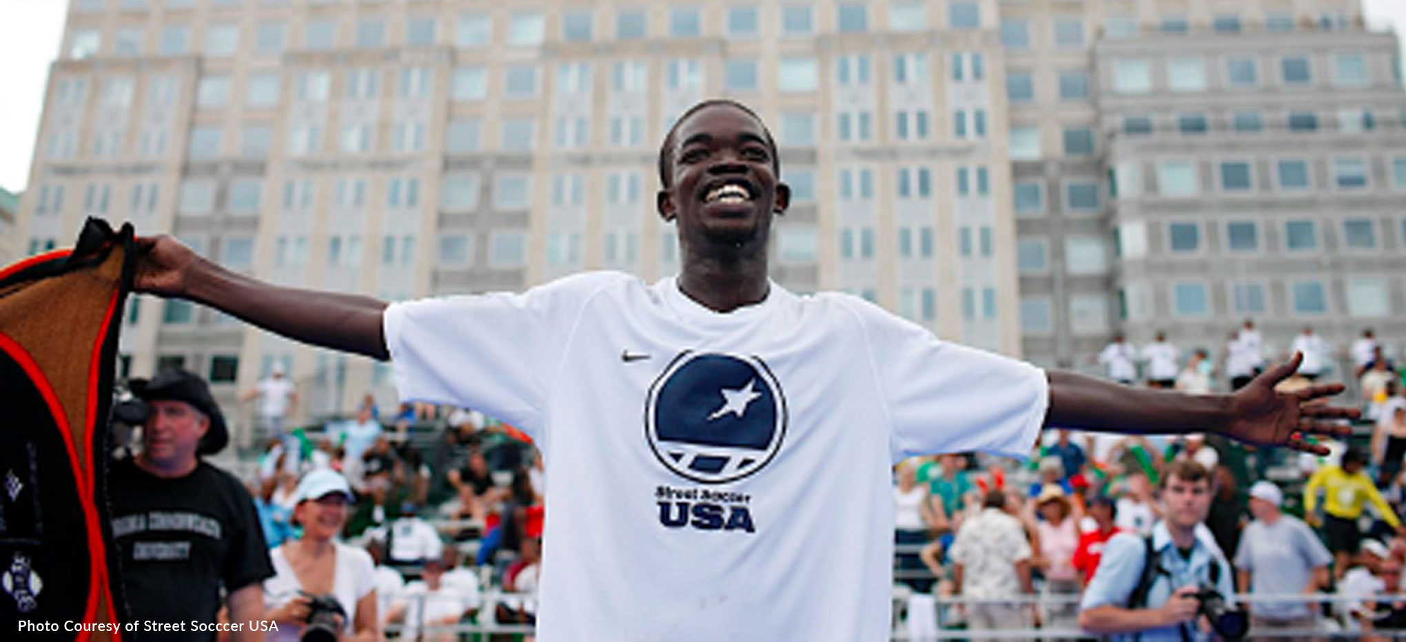A web banner of a black teenage boy wearing a white tshirt with the logo of Street Soccer USA. He stands with his arms open triumphantly, smiling in front of a crowd of people in seats. Several photographers are in the background as well. The lower left of the image contains an accreditation Courtesy of Street Soccer USA.