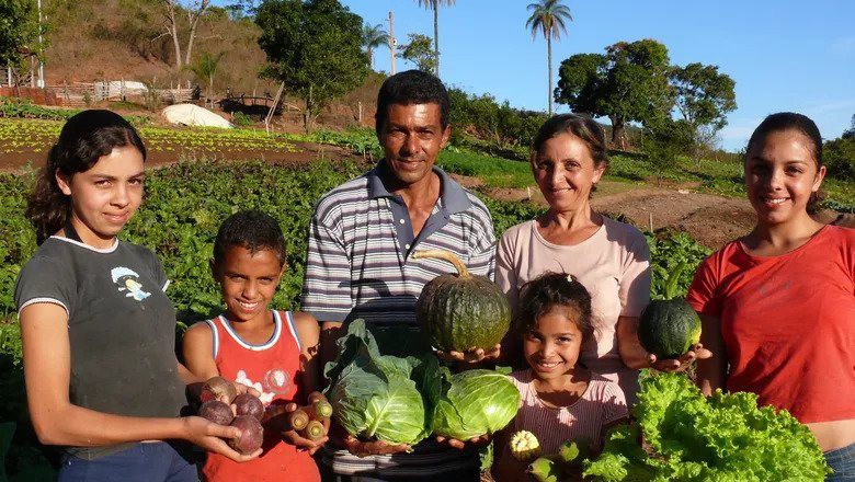 A family of six stands in their field of vegetables holding up crops they've harvested. The mother and father stand at the canter and their daughters around them holding potatoes, leafy greens, and pumpkins.
