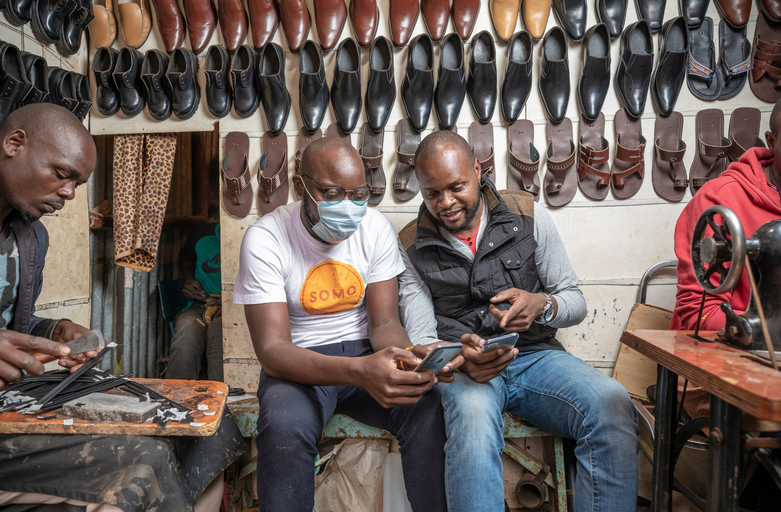 Four men sit in a cobbler shop, two of the men at the edge of the photo are working on pieces for constructing shoes. The two men at the center are speaking with eachother while using their mobile phones.