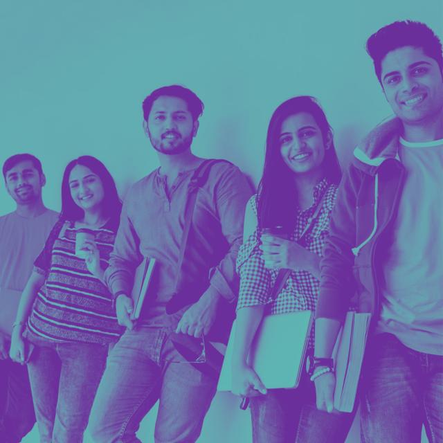 A group of young Indian people leaning casually on a wall, all smiling towards the viewer. Each is holding something associated with school or university.