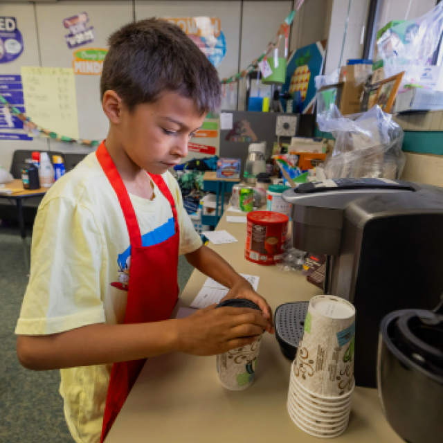 A young boy in a red apron is at his classroms snack station, carefully preparing a hot beverage in a disposable cup.