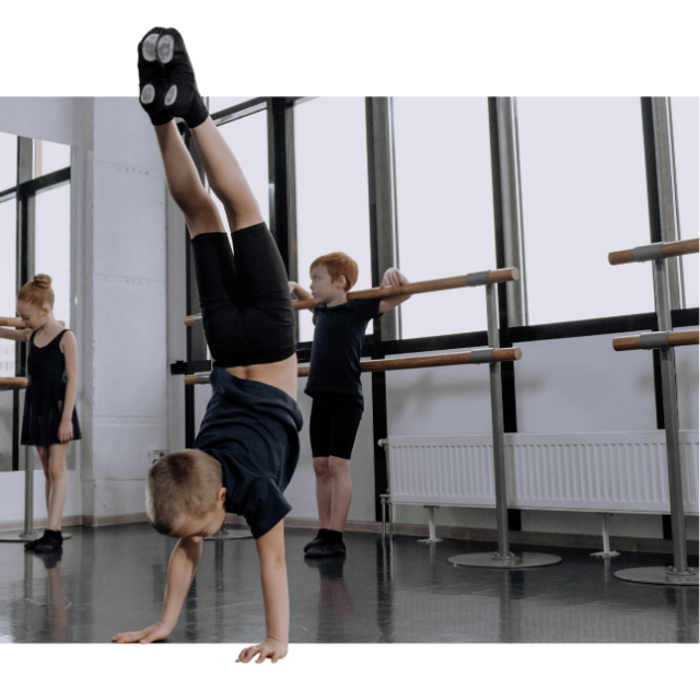 Several children practicing in a ballet studio. A young boy in the foreground is doing a handstand, while a boy and girl in the background stand leaning against the bars.