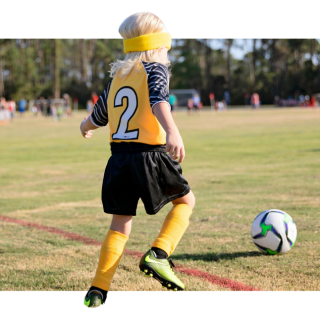 A photo following a child with long blond hair, wearing a yellow number 2 jersey, black shorts, and yellow socks. The child is running to chase after a football.