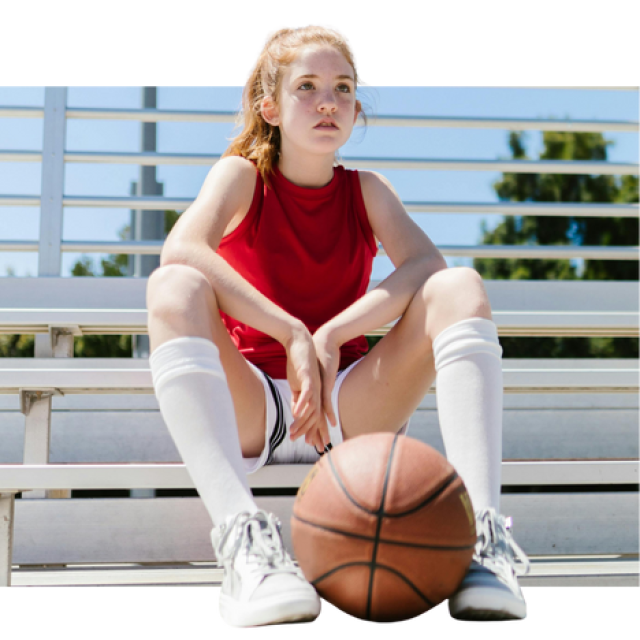 A young girl sits on sports bleachers wearing a red jersey, long white socks and white sneakers. She is holding a basketball between her feet.
