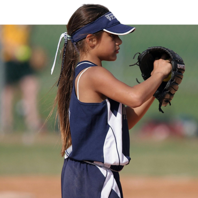 A photo of a girl with tan skin and brown hair, wearing a navy blue jersey and matching visor. Her hand is in her baseball mit as if preparing to throw a ball.