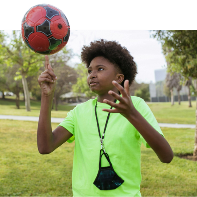 A boy with an afro stands in a park spinning a football on their finger.