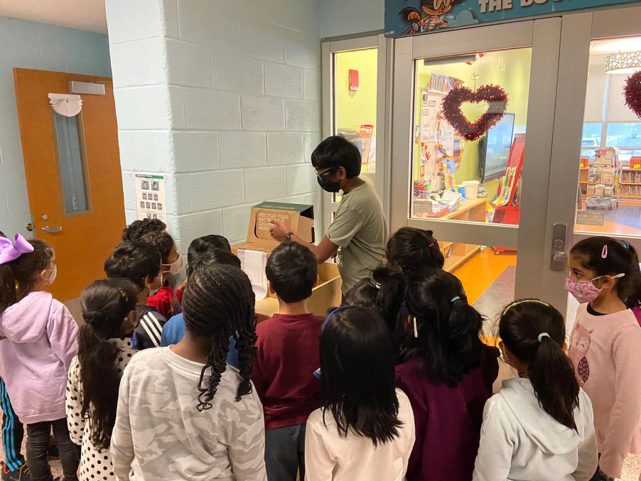 Nihal educating kids about battery recycling at a local school. Nihal is standing in front of a group of students pointing at a recycling bin.