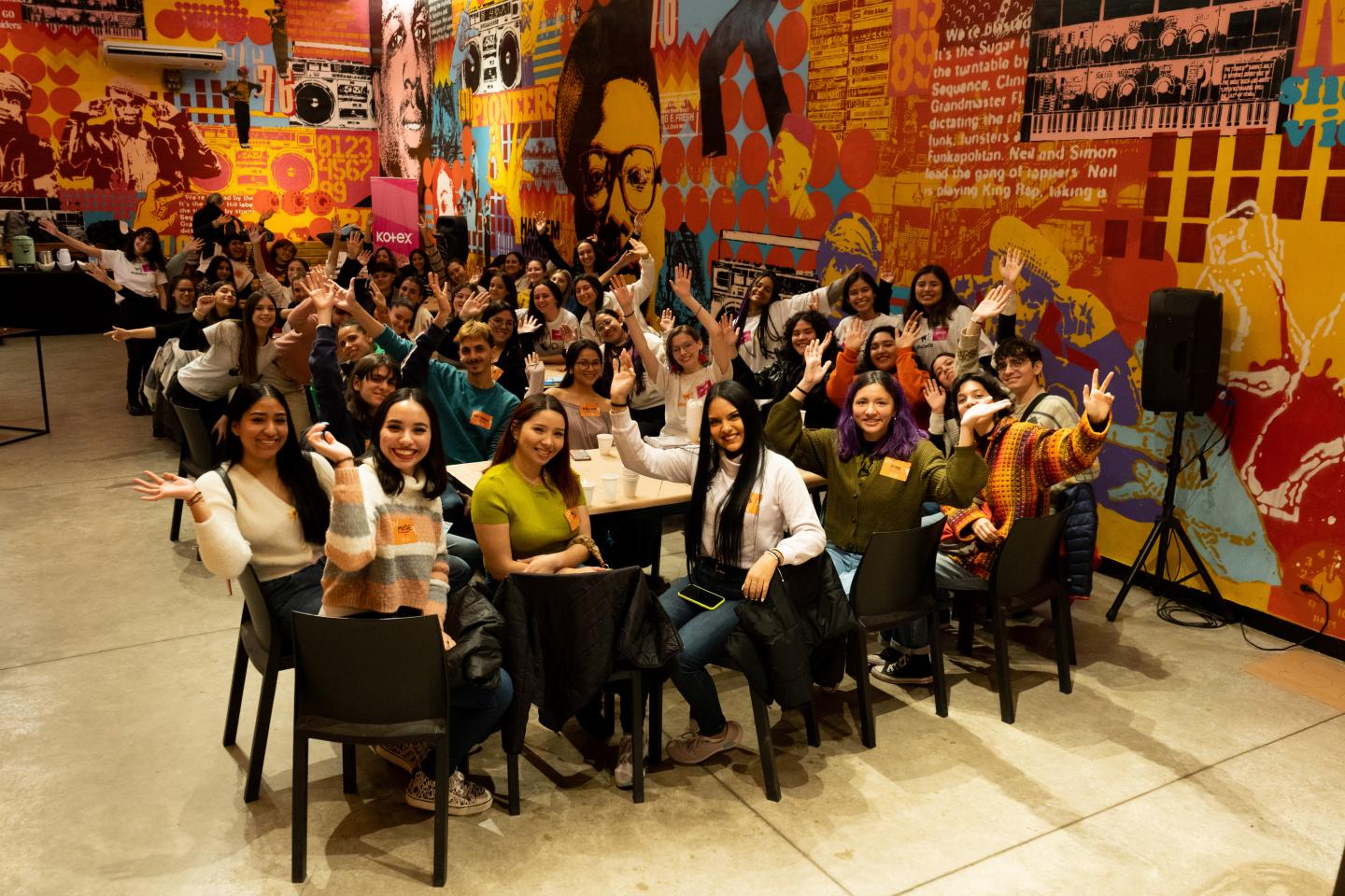 A large group of people sit at tables all posing. The group has raised their arms cheering.