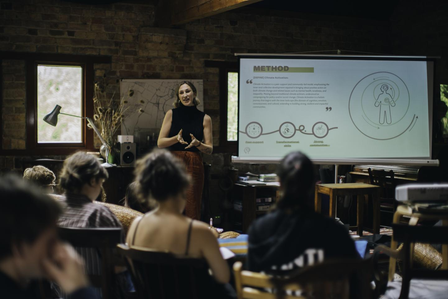 A woman stands at the front of a room nearby to a presentation. Her hands are gesturing as she speaks to an audience of young women in front of her.