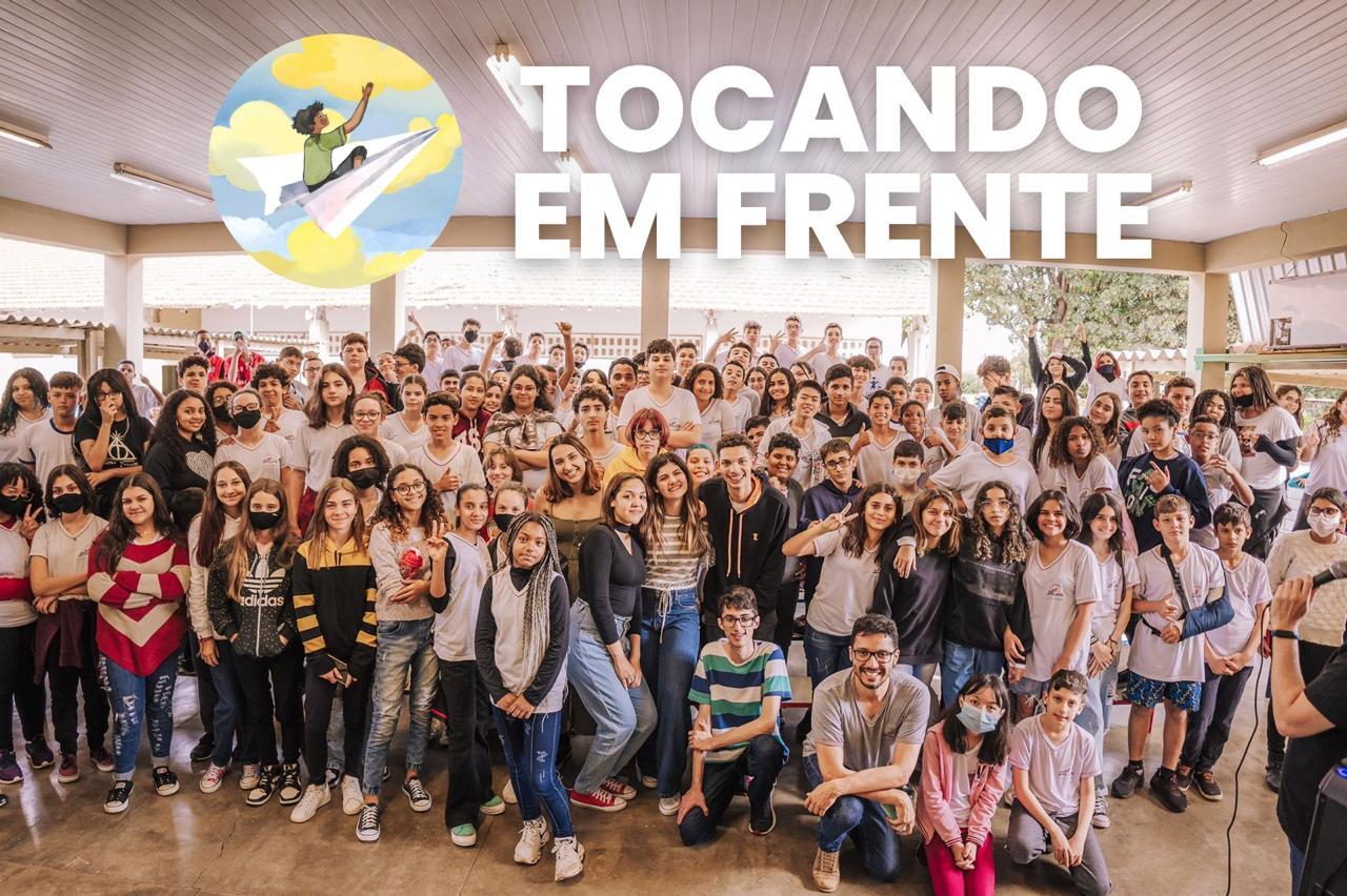A large froup of people pose for a photo. The organization logo reads "Tocando Em Frente" across the top of the photo.