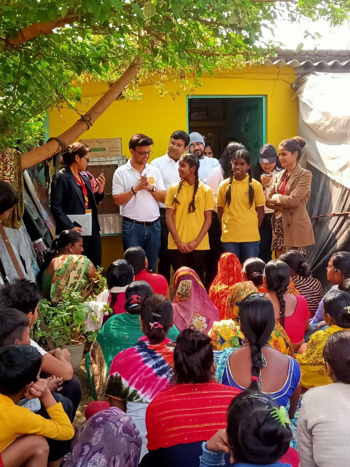 A group of students stand in from of a yellow building, speaking to an audience sitting on the ground in front of them.