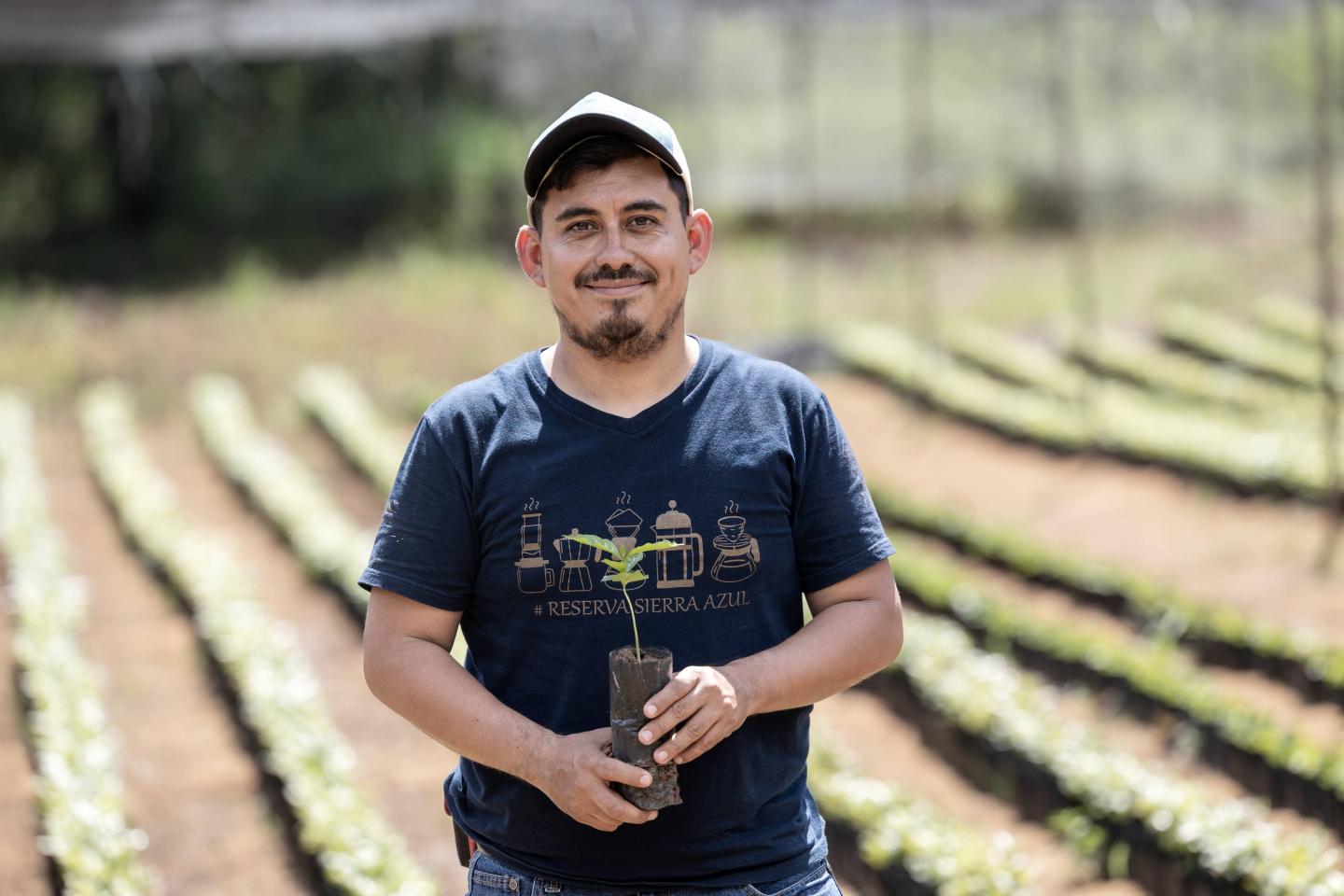 A man holding a sproutling poses in front of rows of vegetation on a farm.