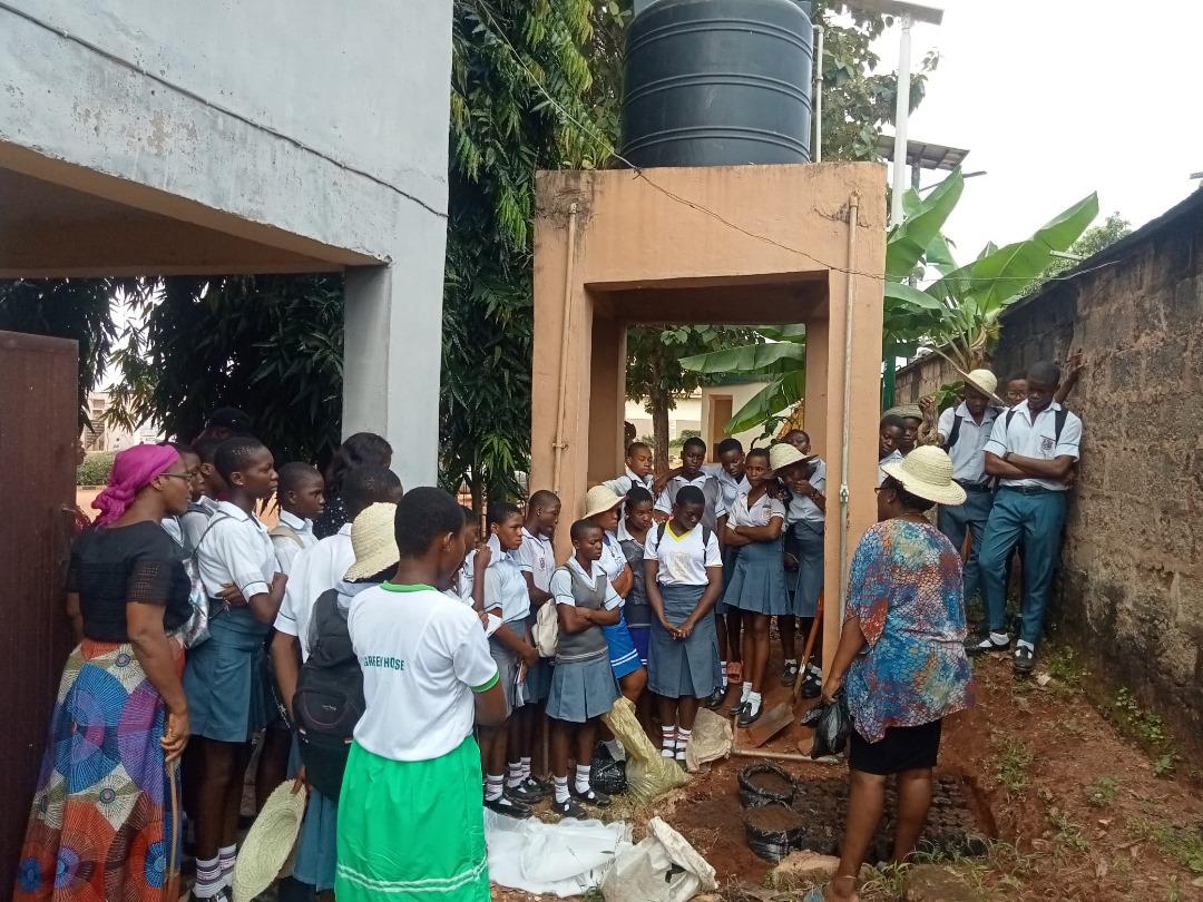 Young people stand in a group outside as they listen to a speaker standing in a shallow dug-out hole.
