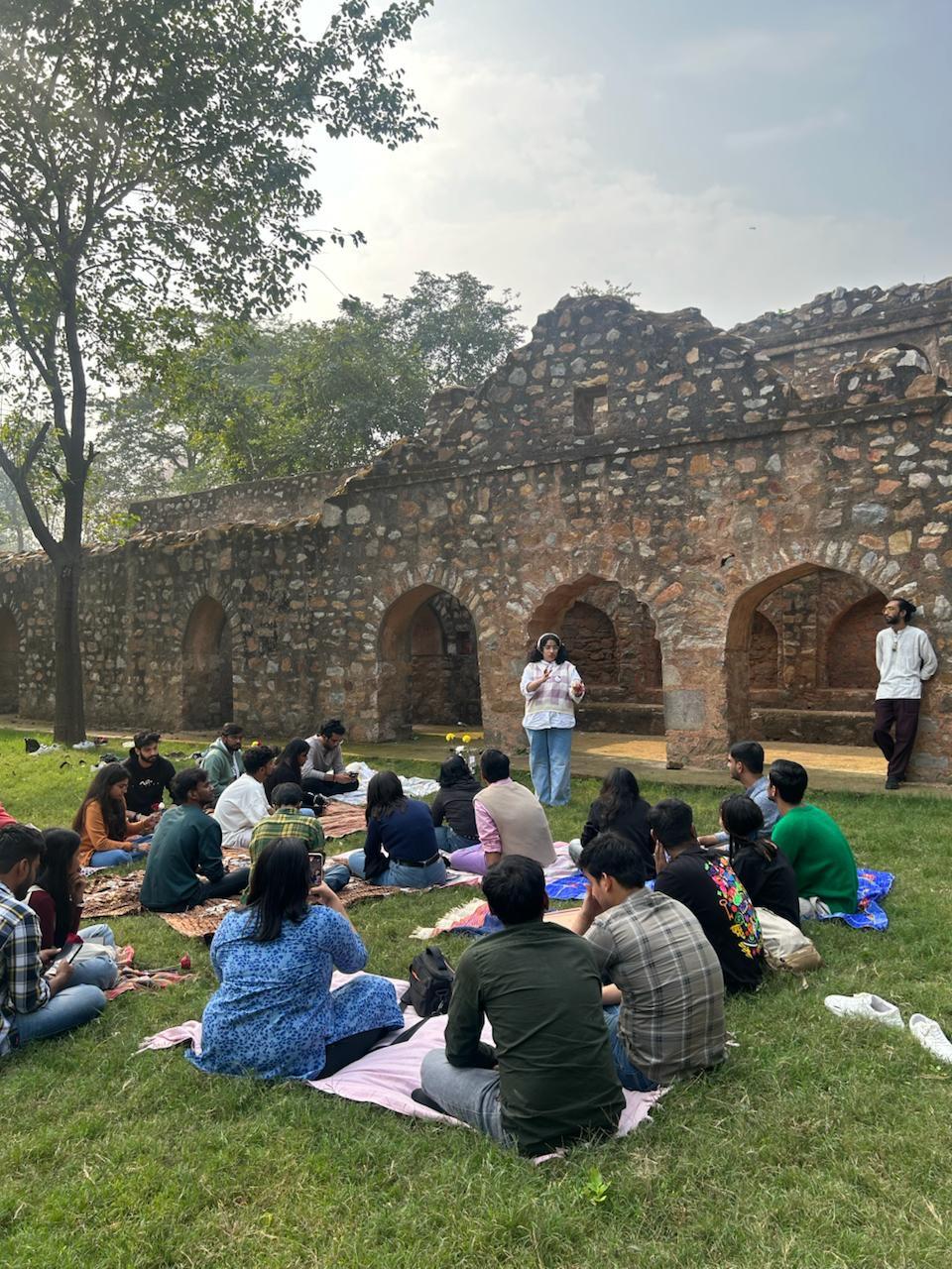 People gathered, sitting on blankets in a grassy field in front of ruins. They are listening to a woman speak in the background.