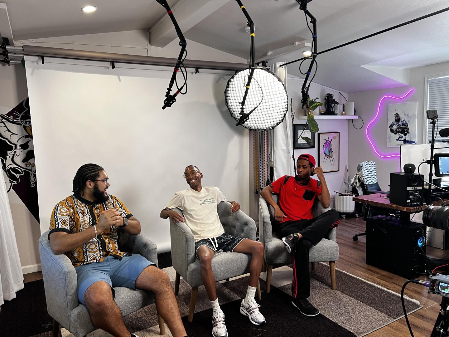 A trio of young black men sit in armchairs next to eachother in a recording studio.