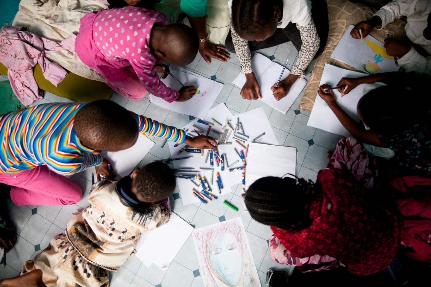 A photo taken from above as several young african children draw with crayons on individual papers.