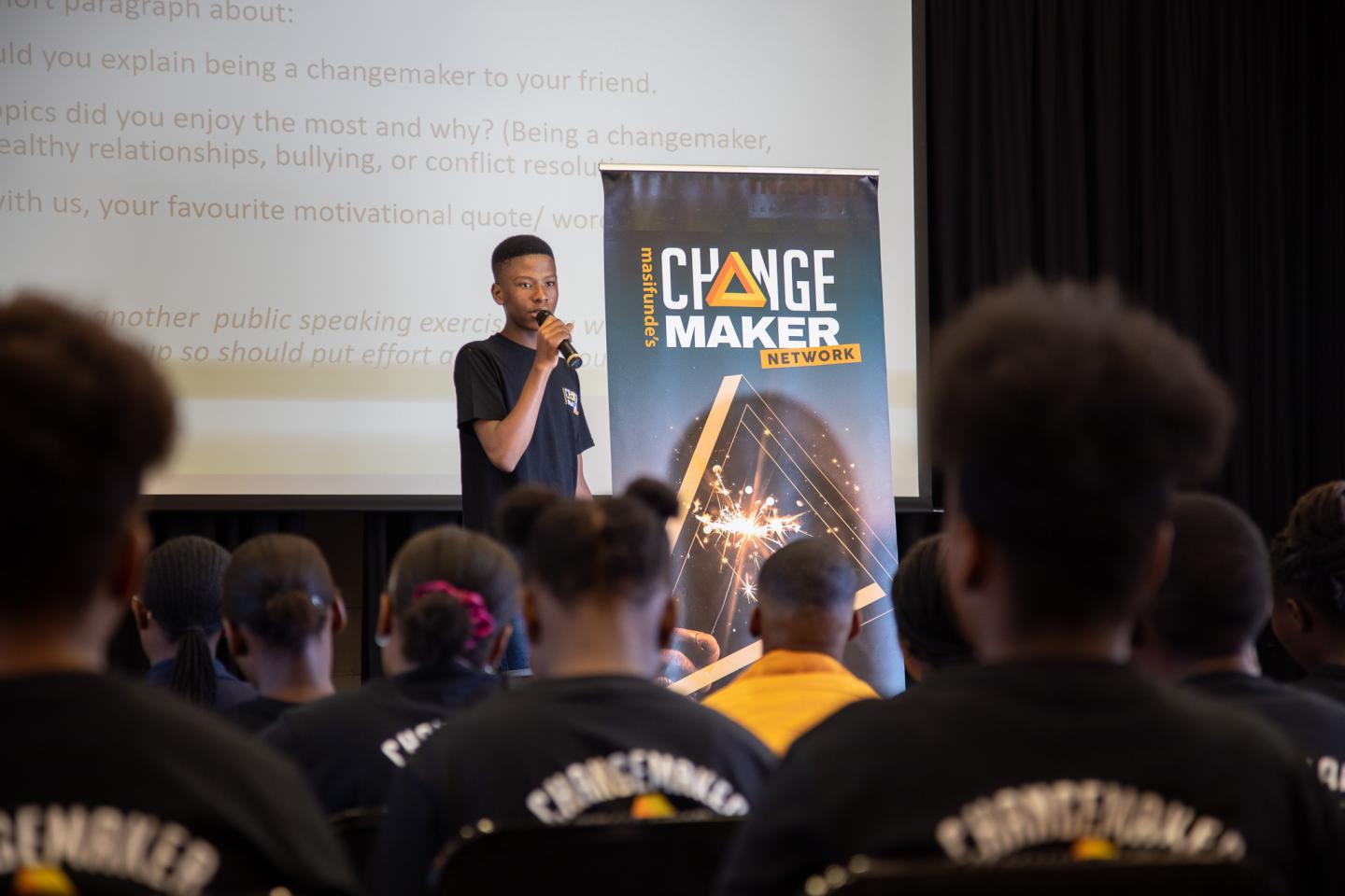 A young man speaks on a stage to a large audience. He is standing next to a banner that reads "Changemaker". Members of the audience are all wearing shirts with the same logo on the back.