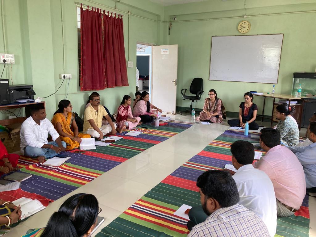 A group of people sit in a circle on the floor in what looks like a classroom. They each have books in front of them as they listen to a person at the front of the room speak.