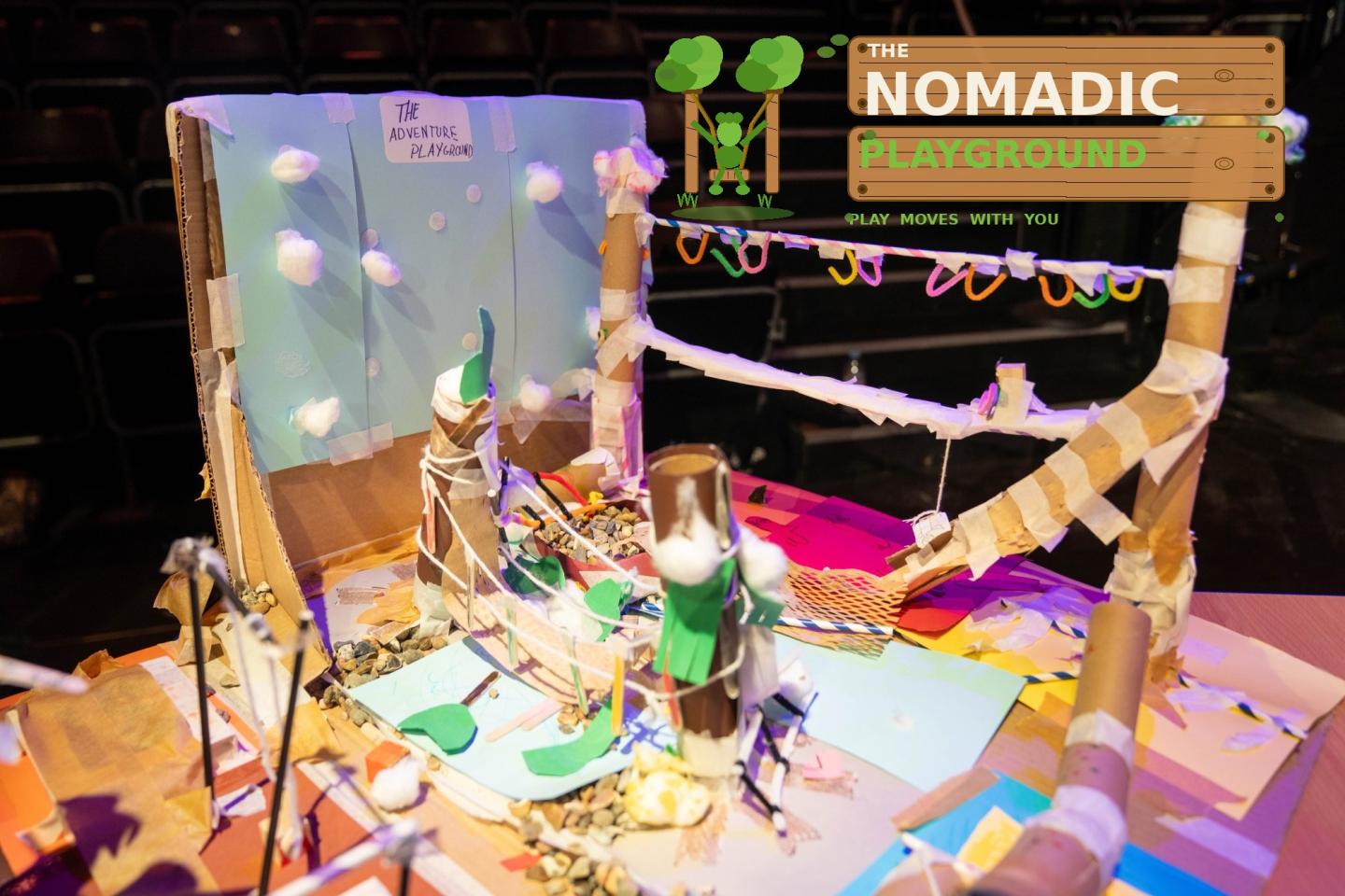 A photo of a diorama of a playground constructed from recylced and craft materials, including a bridge made of popsicle sticks and gymnastic equipment made from straws. There is a logo that reads "The Nomadic Playground Play Moves With You" in the top right corner.