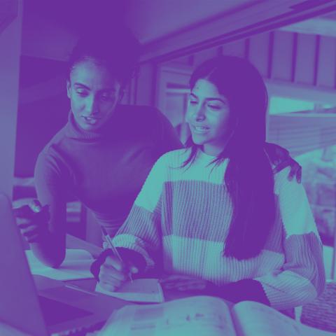 Two Indian women working at a laptop computer. One is pointing at the screen while the other is taking notes in a notebook.