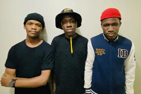 Three young black men stand against a blank white wall wearing casual clothing. One wears a bucket hat, one a black beanie and black shirt, and the last wears a red beanie with a letterman jacket.