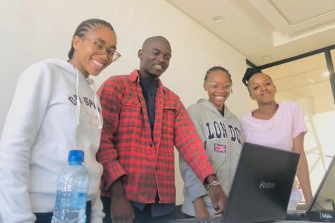 Four young people, two boys and two girls, stand at a desk looking at a laptop.