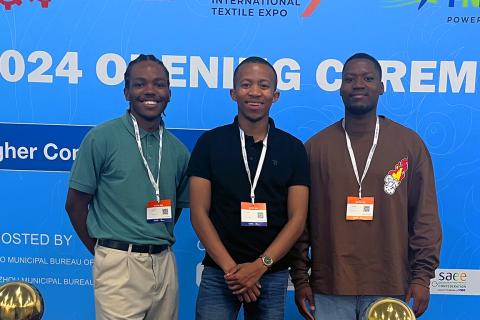 Three young men stand shoulder to shoulder in front of a step and repeat.