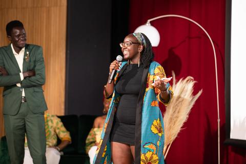 A young woman wearing a coat featuring an teal and yellow, floral, African print speaks into a microphone at a stage to an unseen audience.
