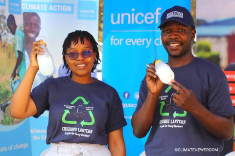 Two young students wearing branded shirts that read "Chicubhu Lantern A Light for Every Student", stand in front of Unicef banners. They are posing with the invention, a small sustainably-made lantern.